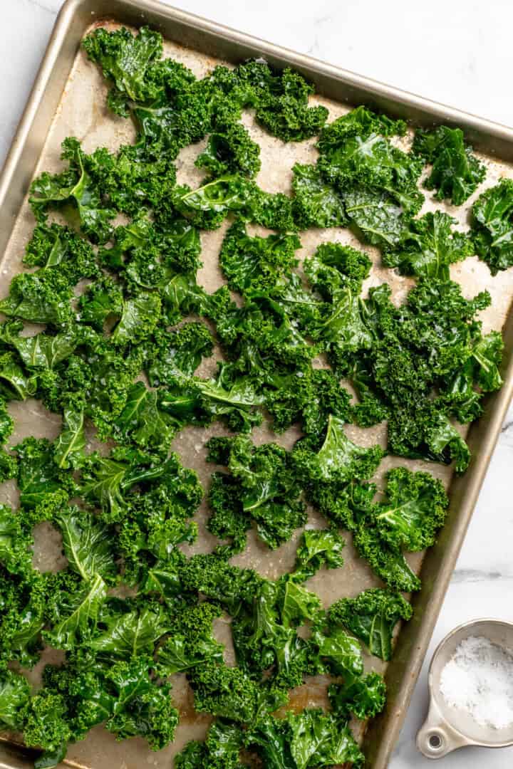 Whether you season them with sea salt, black pepper, or vegan Parmesan, these crispy baked kale chips are an easy snack ready in minutes! Kale leaves tossed in oil on sheet pan.