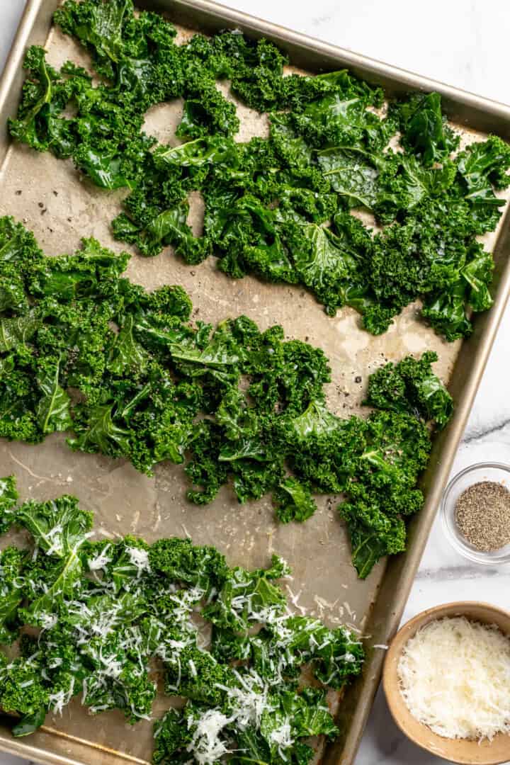 Whether you season them with sea salt, black pepper, or vegan Parmesan, these crispy baked kale chips are an easy snack ready in minutes! Kale on sheet pan, divided into three sections with sea salt, vegan parmesan, and black pepper.