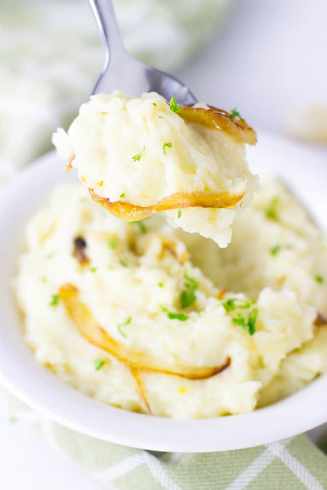 Spoon with garlic mash potatoes in front of a bowl of mash potatoes. 