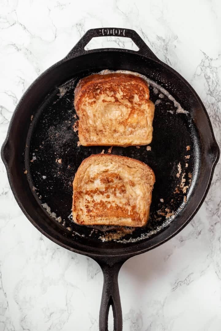 Two slices of eggnog French toast cooking in cast iron skillet.