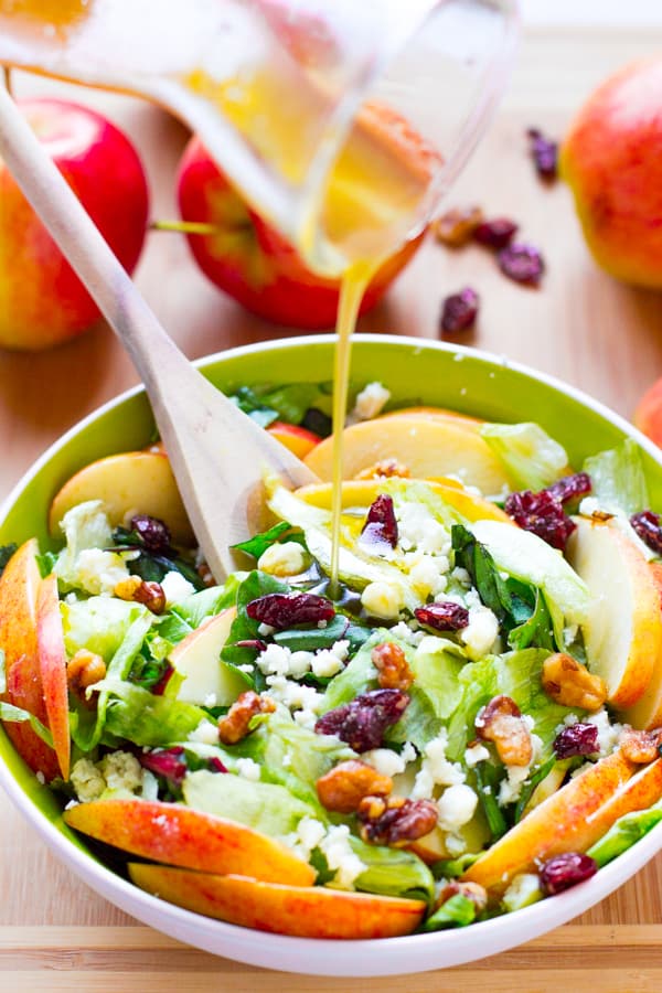 Dressing being poured onto apple and blue cheese salad in green bowl. 