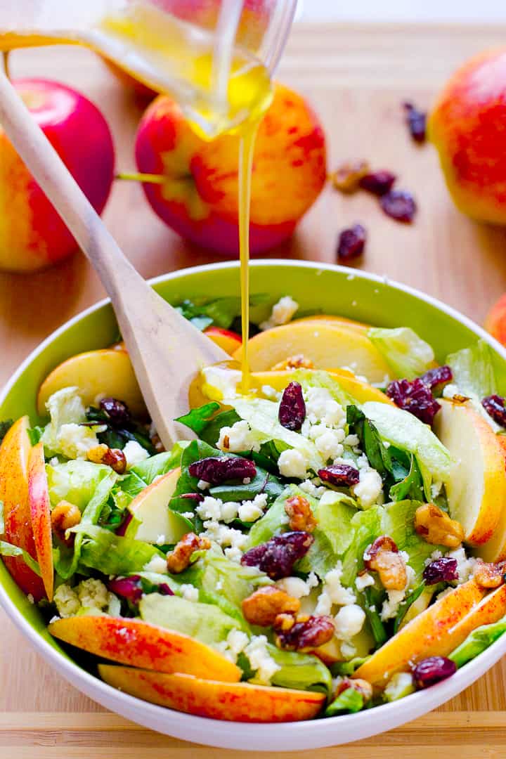 Pouring dressing on apple, candied walnuts and blue cheese salad in a green and white bowl. 