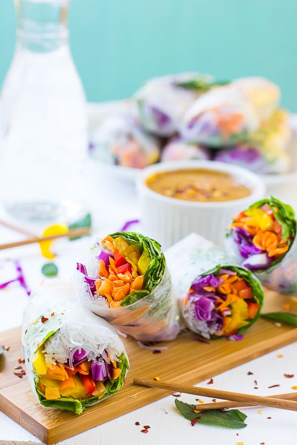 Four fresh spring rolls on wooden board with stack of rolls on plate in background