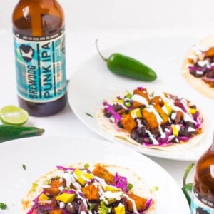 Black bean and sweet potato tacos on white plates with two bottles of beer.