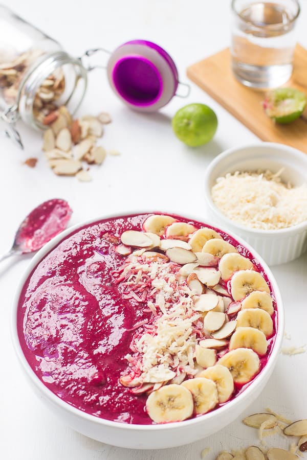 Banana berry beet smoothie bowl on a white table top with ingredients around it. 