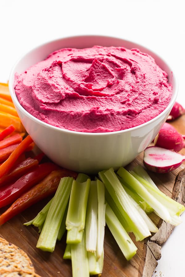 Side shot of beet hummus in a white bowl with raw vegetables on the side. 