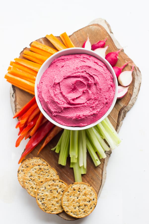 Top down shot of beet hummus in a white bowl with raw vegetables and crackers. 