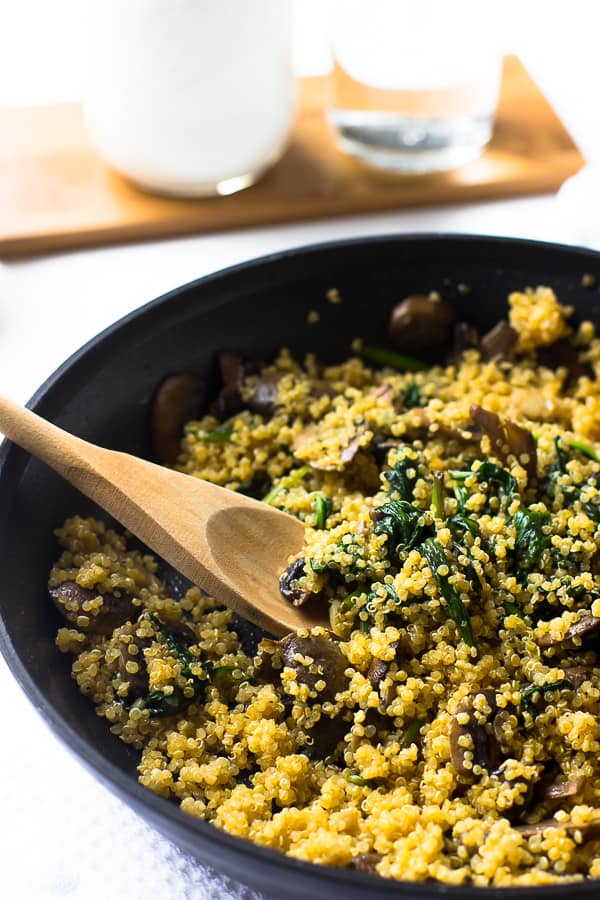 Spinach and mushroom quinoa in a black skillet being stirred with a wooden spoon. 