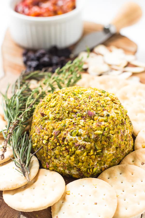 Pistachio crusted vegan cheese ball on a cheese board with crackers and a sprig of rosemary.