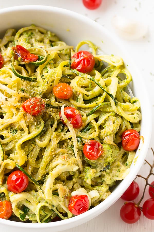 Pesto zucchini noodles with burst cherry tomatoes in a white bowl with cherry tomatoes on the side. 