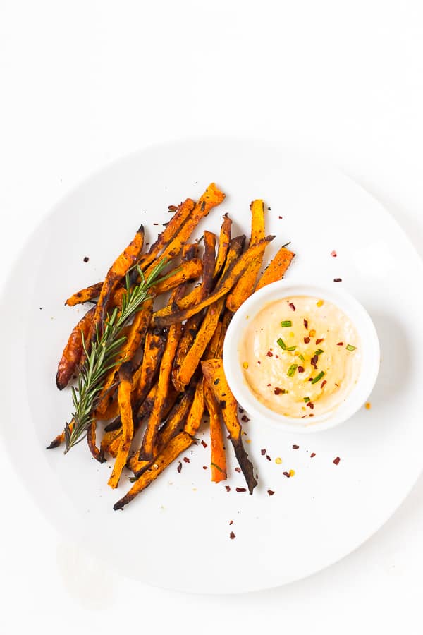 Overhead shot of crispy baked carrot fries on a white backdrop with harissa tahini dip in a ramekin.