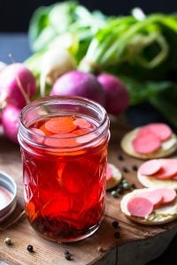 Pickle radishes in a glass jar on a board with radishes in the background.