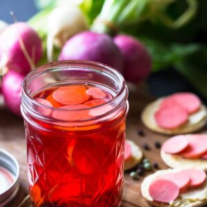 Pickle radishes in a glass jar on a board with radishes in the background.