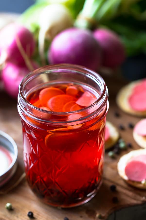 Pickled radishes in a glass jar surrounded by raw radishes.