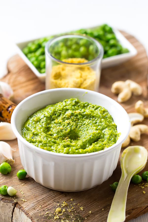 Bright green creamy cashew and pea pesto in a white ramekin surrounded by peas and nuts. 