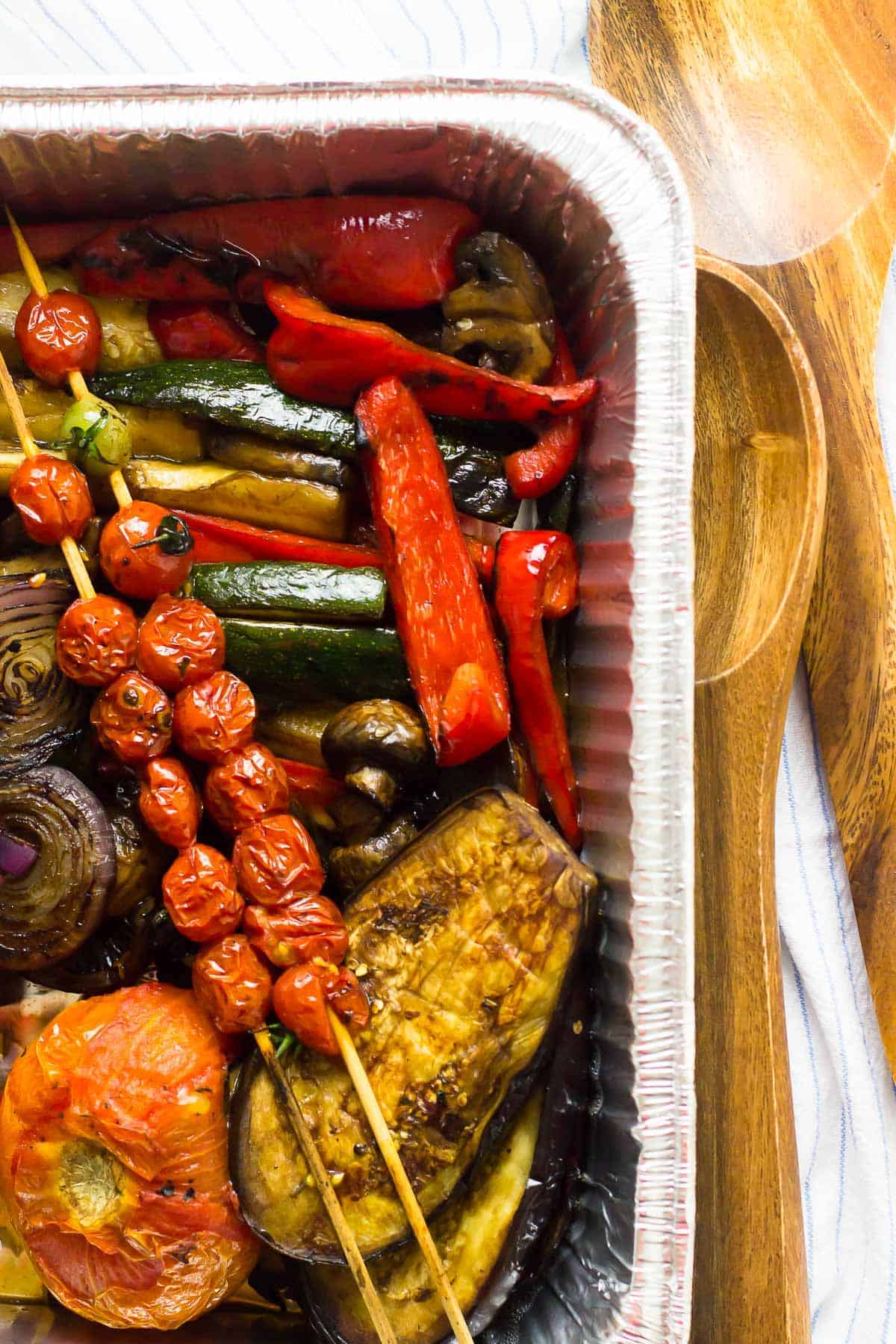 Grilled vegetables in a foil container with wooden servers on the side.