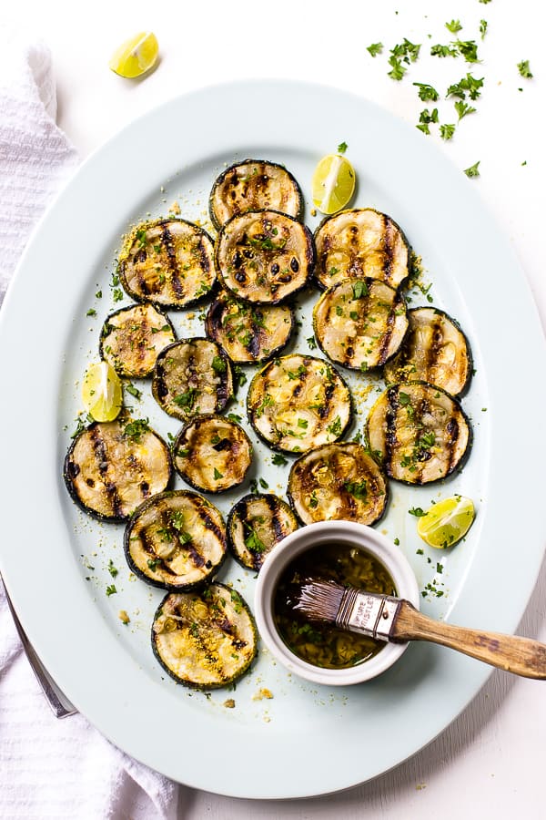 Overhead view of grilled zucchini slices on platter with small bowl of marinade and brush