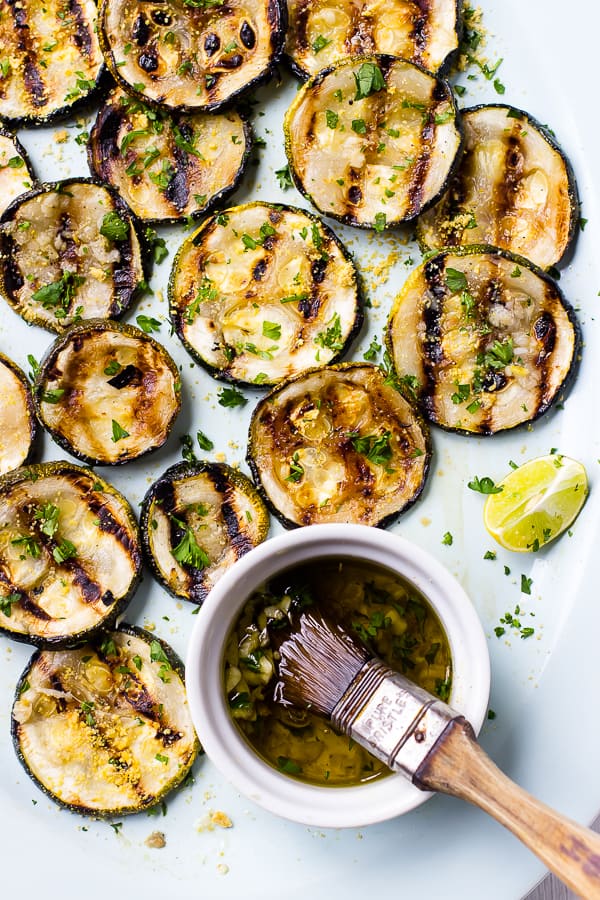Closeup view of lemon garlic grilled zucchini slices on platter with small bowl of marinade and brush