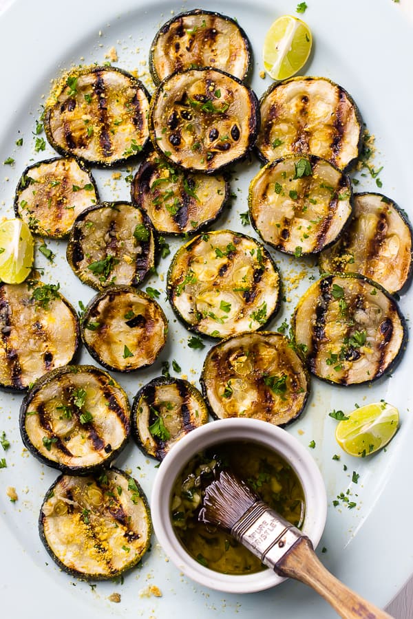 Overhead view of grilled zucchini slices on platter with small bowl of marinade and brush