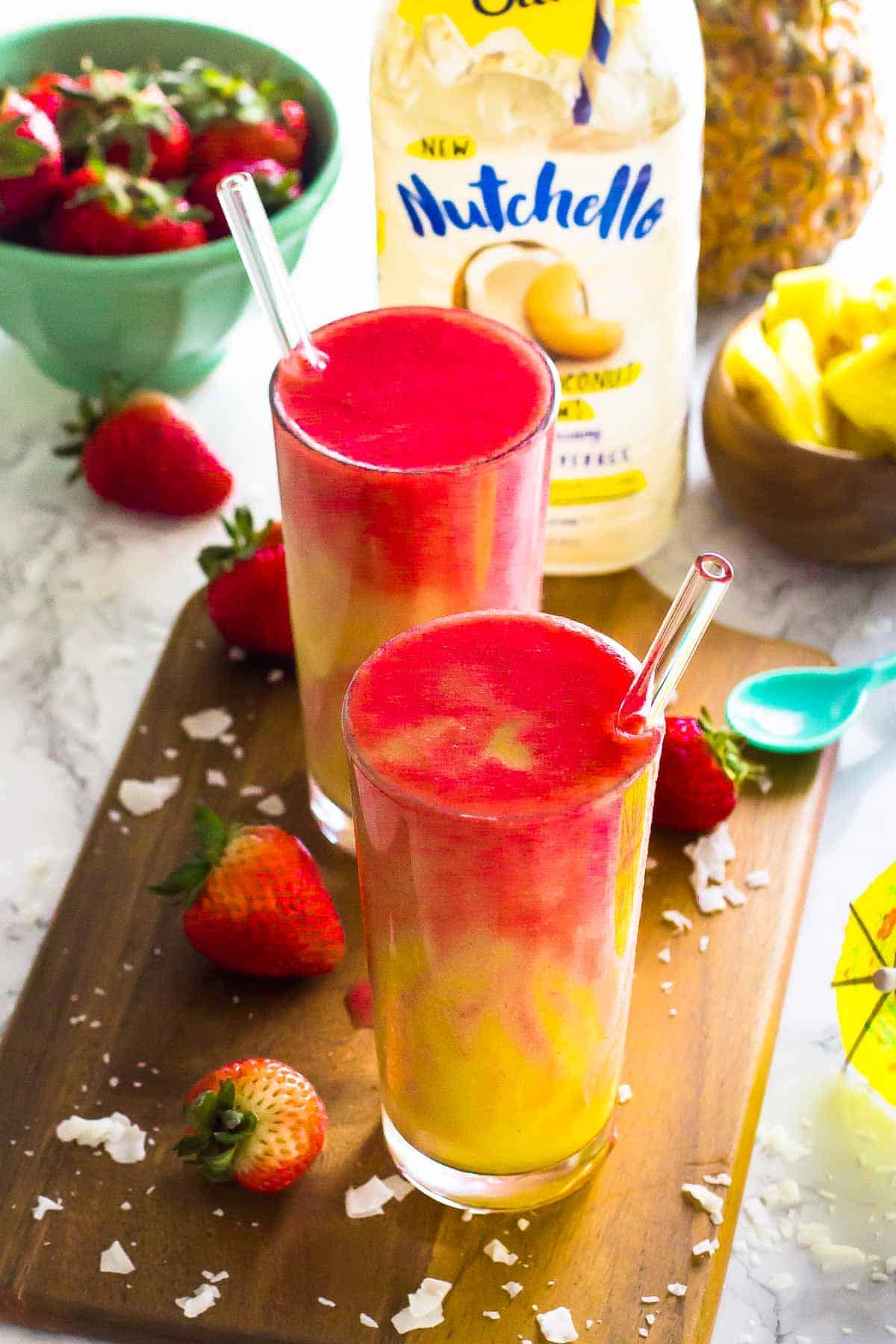 Strawberry pineapple coconut smoothies in two tall glasses on a wood board. 