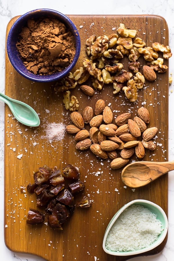 Ingredients to make no bake brownie energy bites on a cutting board, almonds coconut flakes, dates, coco powder, salt and walnuts.