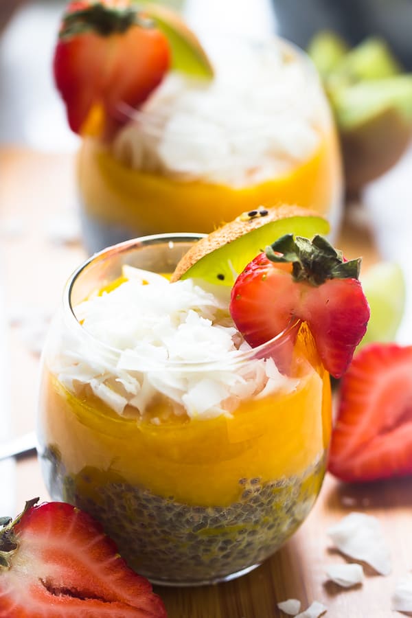 A mango coconut chia seed pudding in a wine glass, garnished with a slice of strawberry and kiwi, with another pudding in the backgrounds, and strawberrys and coconut flakes on the table