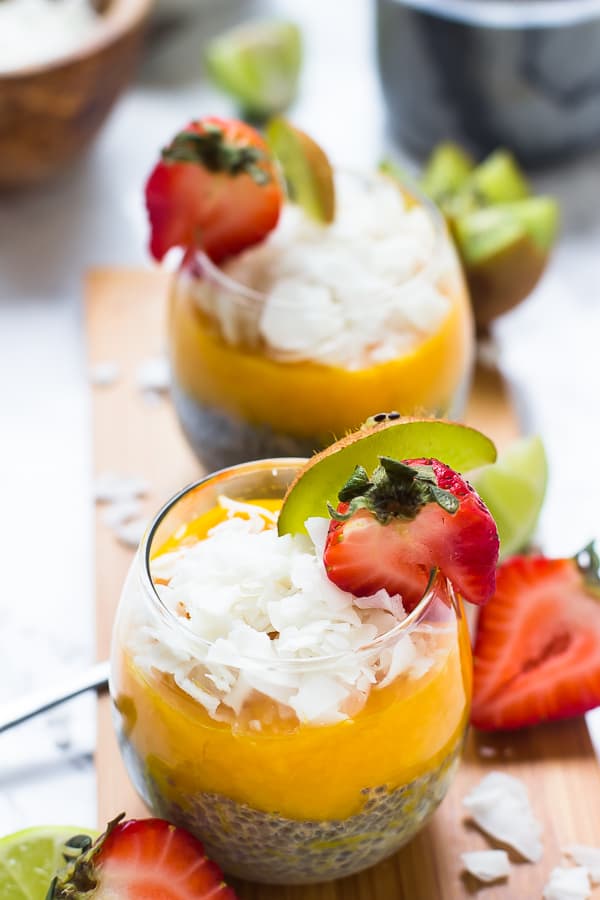 A mango coconut chia seed pudding in a wine glass, garnished with a strawberry slice and a kiwi slice, with another pudding in the background, and strawberries and kiwis on the table 