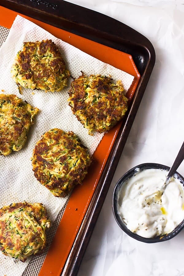 Top down shot of vegan zucchini fritters on a baking tray with sauce on the side.