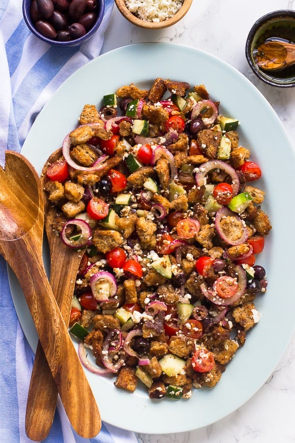 Top down shot of easy greek panzanella salad on a white plate with wooden spoons on the side. 