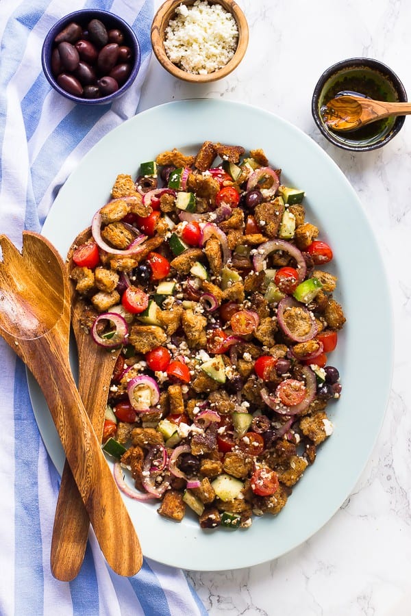 Top down shot of greek panzanella salad on a white dish with a bowls of olives, olive oil, and vegan feta cheese on the side. 