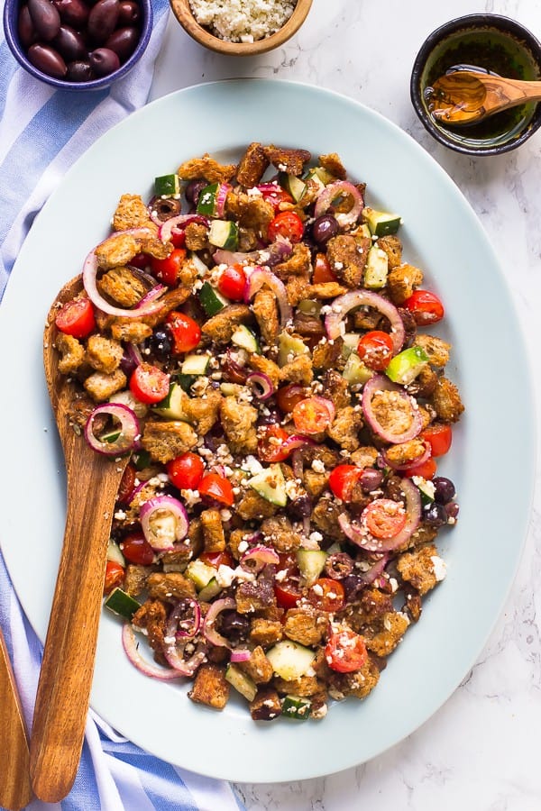 Overhead shot of greek panzanella salad with a wooden spoon, next to a bowl of olives, feta, and salad dressing.