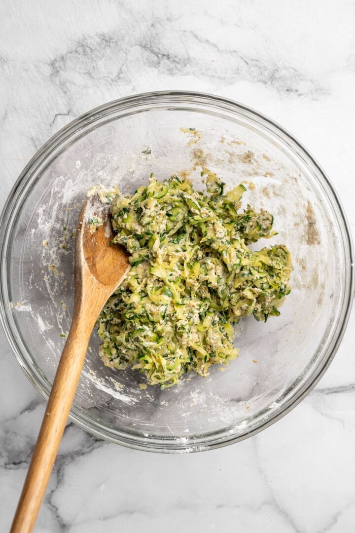 Made with coconut flour, these crispy zucchini fritters are gluten-free and vegan! They're easy to make, perfect for a side or appetizer. Zucchini fritter mixture in glass bowl with wooden spoon.
