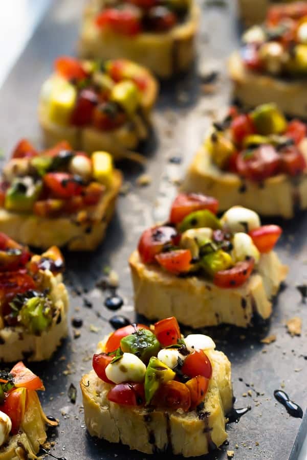 Two rows of avocado caprese bruschetta topped with a balsamic reduction on a grey slate. 