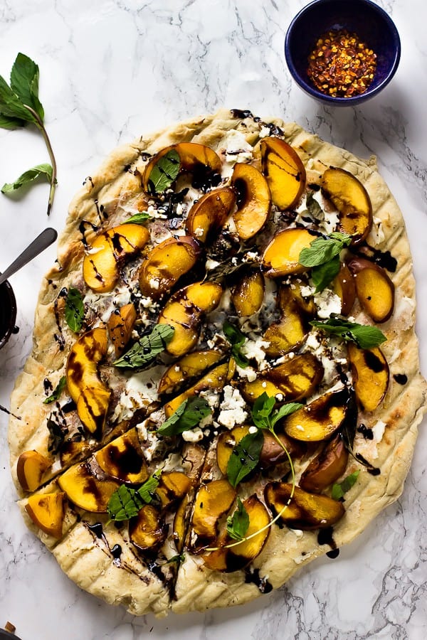 Overhead shot of grilled peach, basil and goat cheese pizza on a marble counter with a bowl of chili flakes on the side. 