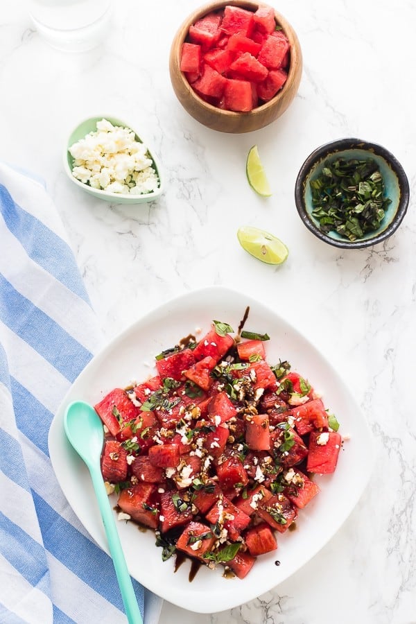 Overhead-Aufnahme von Wassermelonen-Feta-Salat auf einem weißen Teller mit einer Schüssel gewürfelter Wassermelone an der Seite.