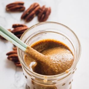 A spoon scooping out pecan butter from a glass jar.