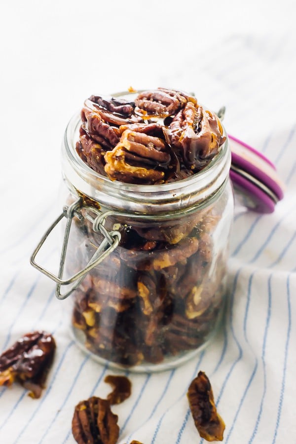 A glass jar full of candied pecans with some pecans around it. 