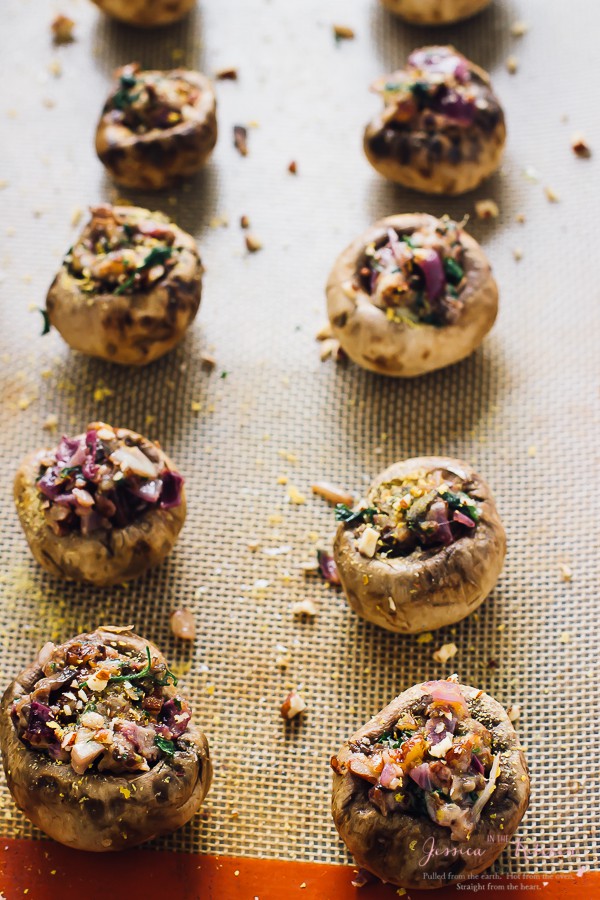 Two rows of raw stuffed mushrooms with coconut bacon on a silpat, with lots of chopped nuts spread around