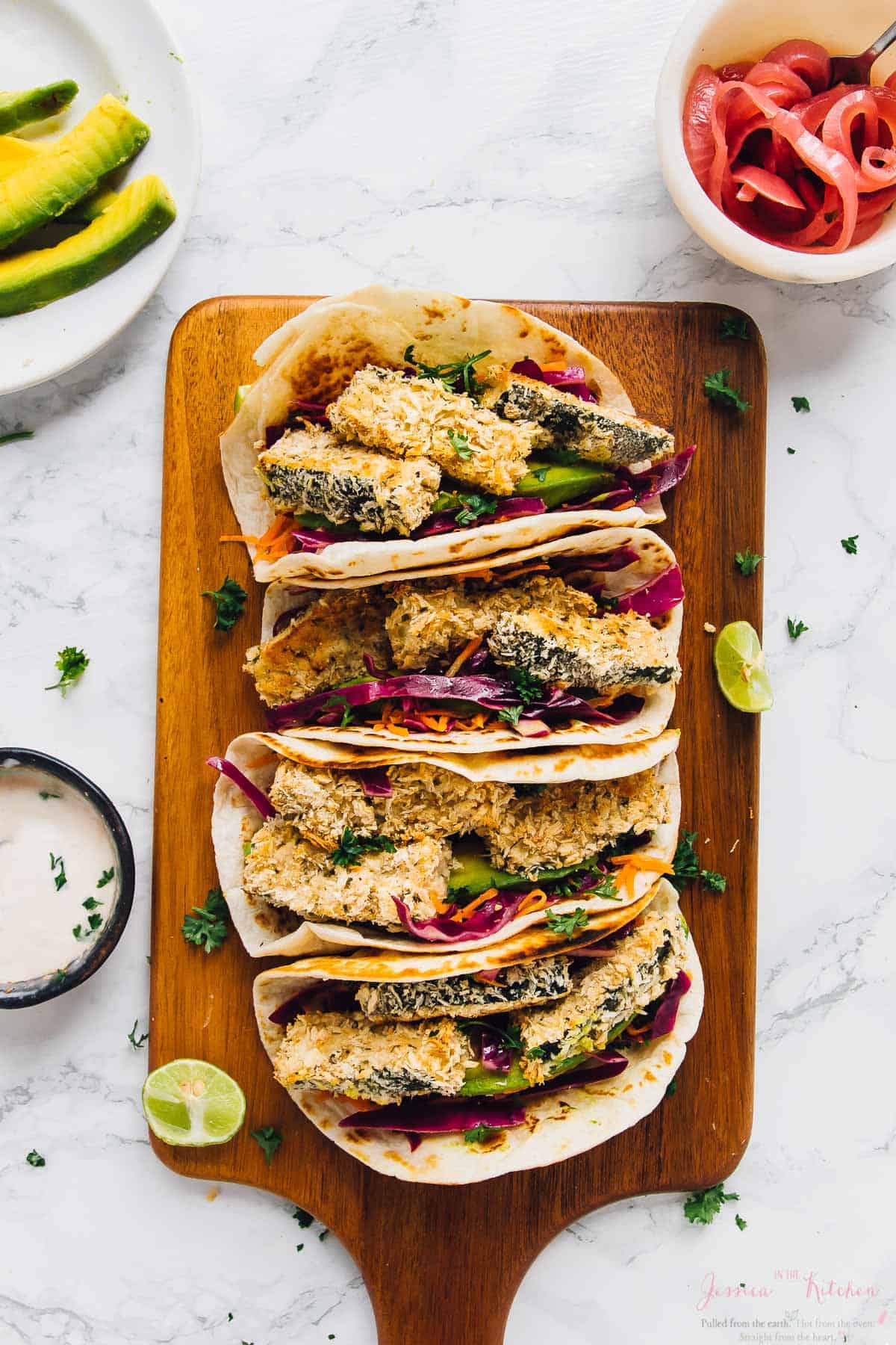 Overhead view of 4 crispy zucchini tacos lined up on a wood board with garnishes in background