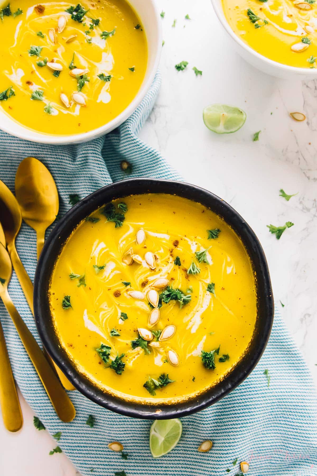 Overhead shot of vibrant yellow vegan butternut squash soup in three bowls with gold spoons on the side. 