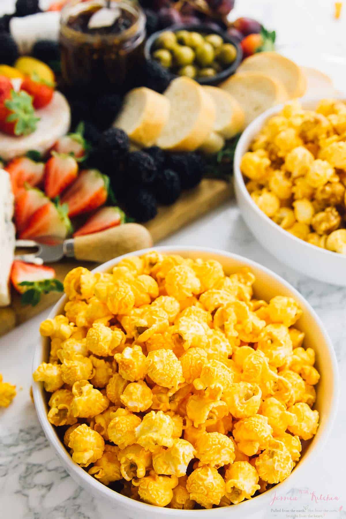 A bowl of cheese popcorn in a bowl with fruit and bread in the background. 