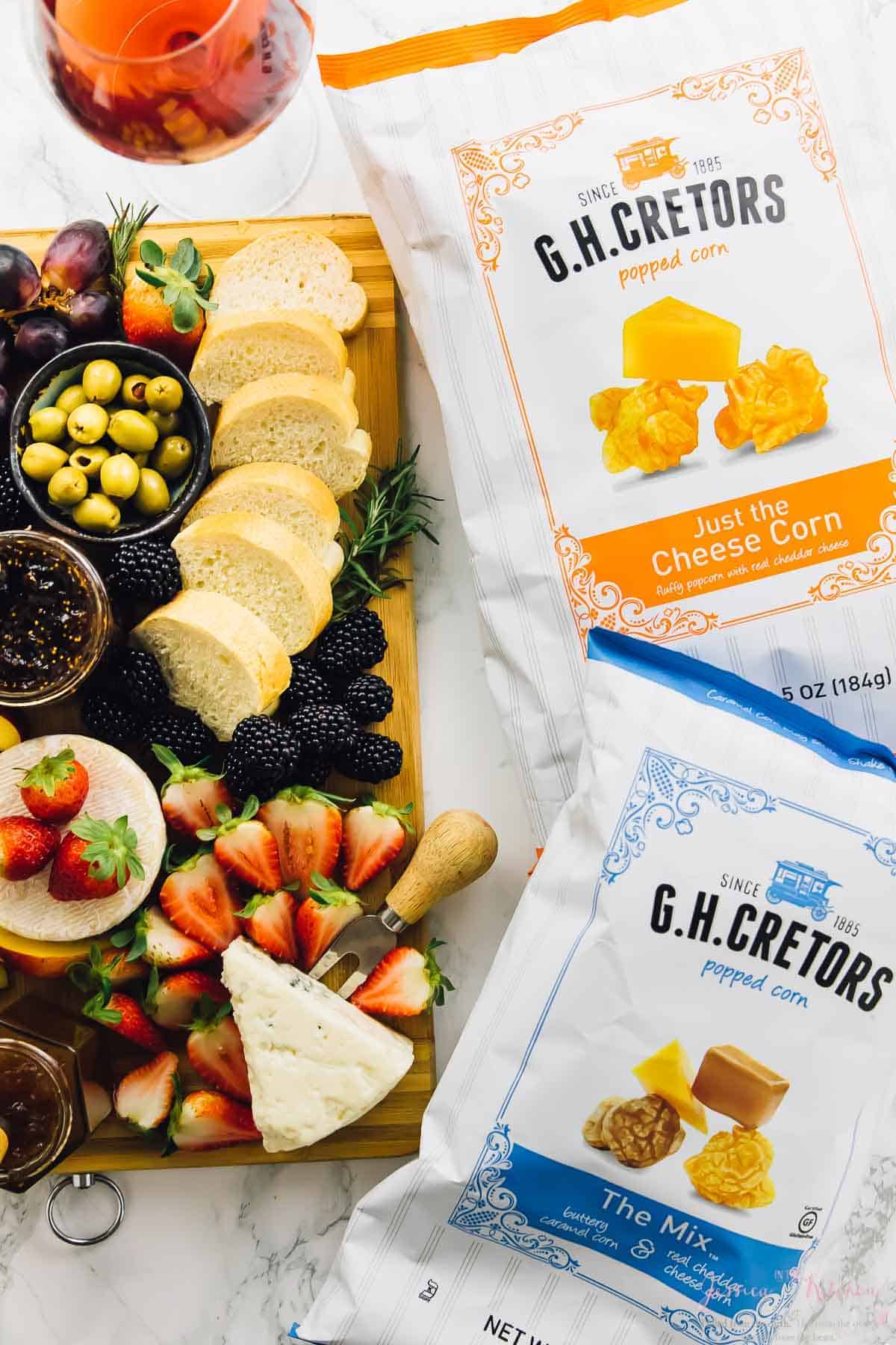 A wood board with fruit, cheese and bread next to bags of popcorn. 