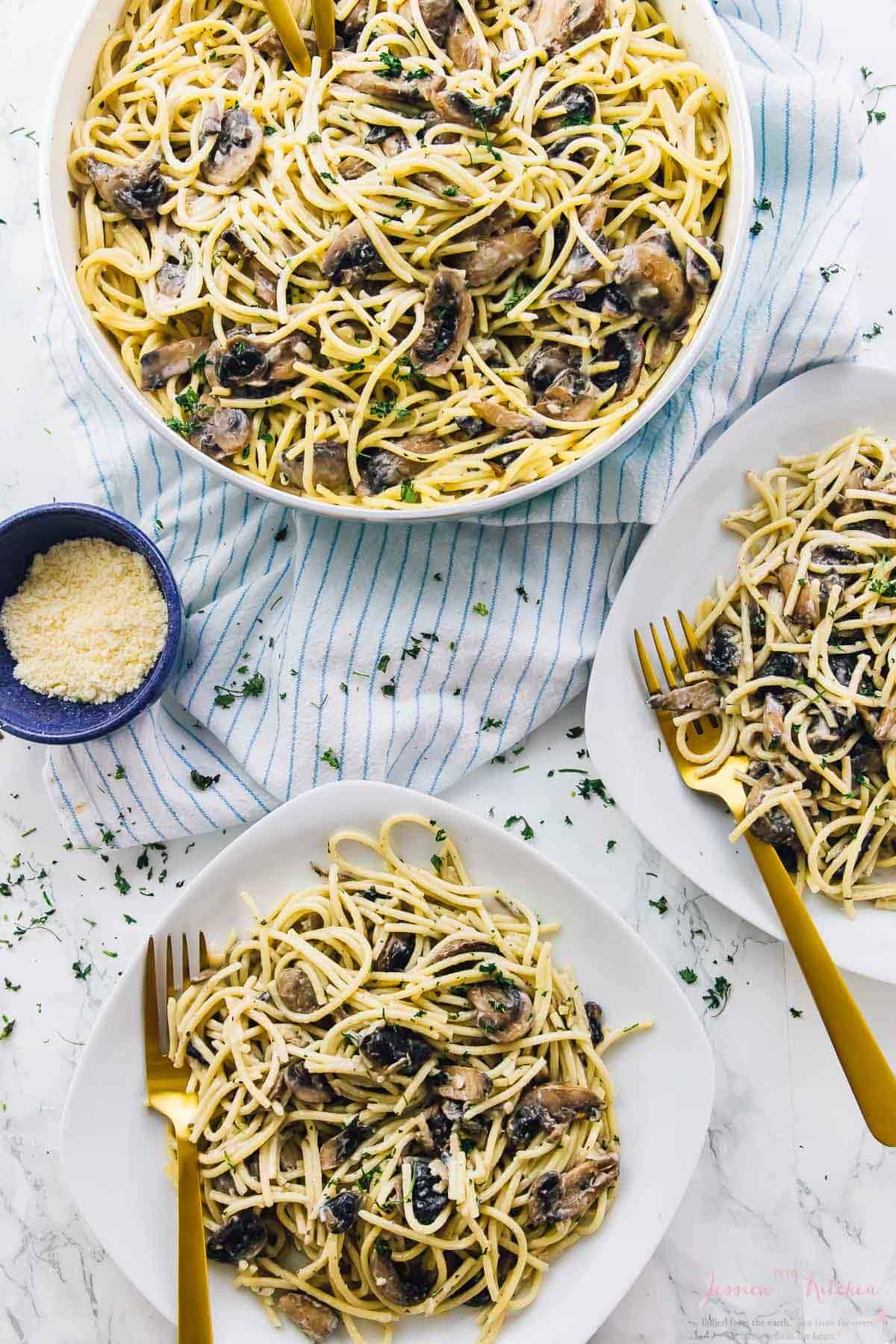 Two plates of mushroom vegan alfredo pasta with gold forks and a bowl of parmesan cheese