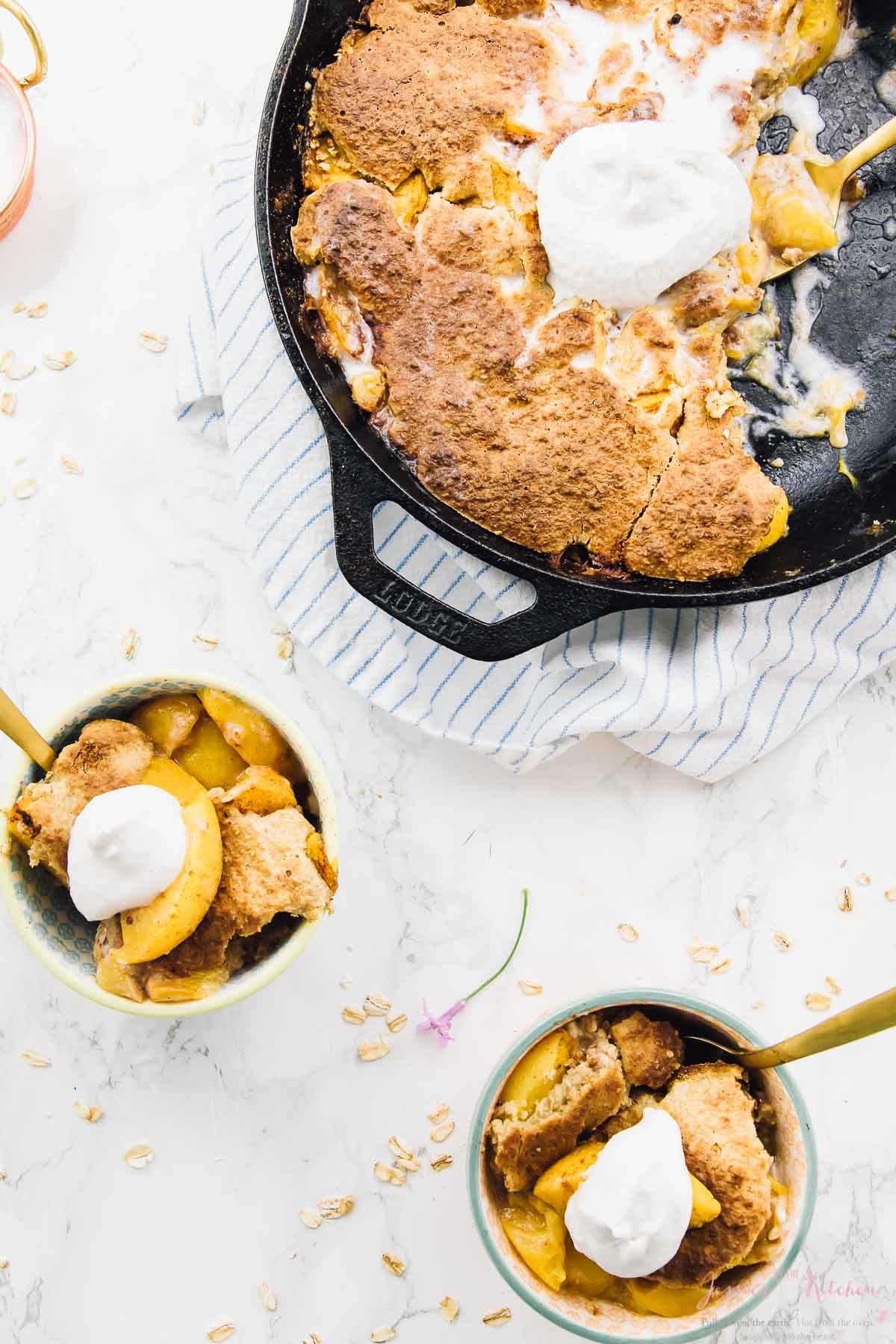 Overhead shot of peach cobbler in a black skillet with cream on top, with two portions in small bowls