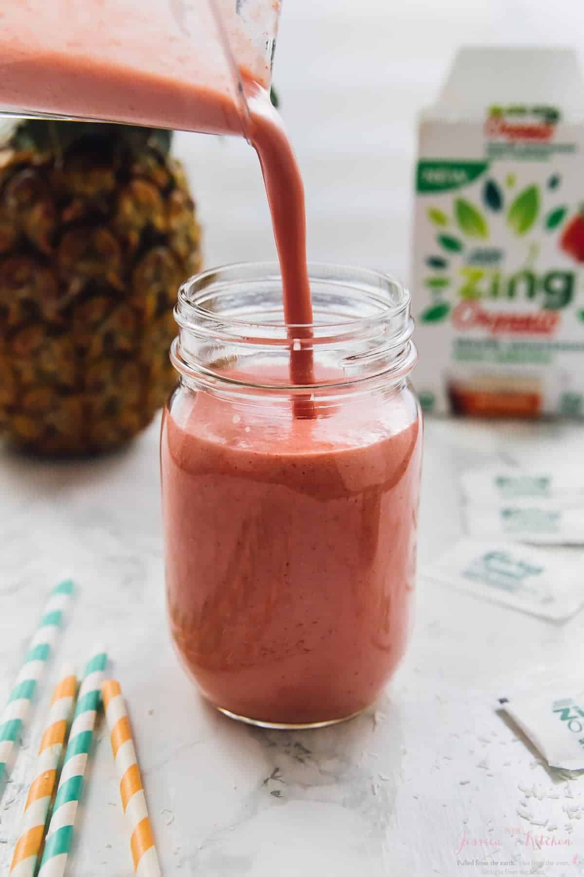 Pouring strawberry smoothie into a mason jar with a pineapple in the background. 