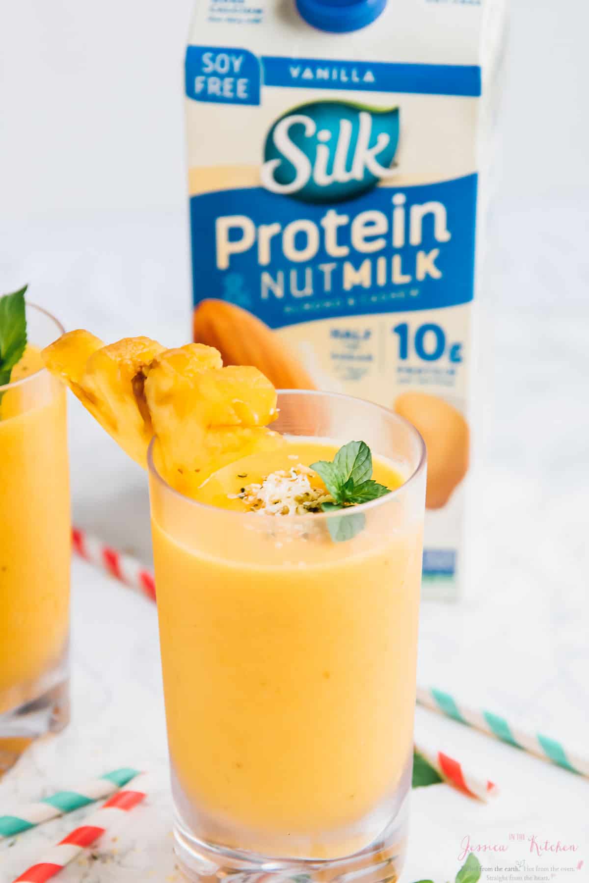 A pineapple peach smoothie in a glass garnished with a pineapple slice and a carton of nut milk in the background. 