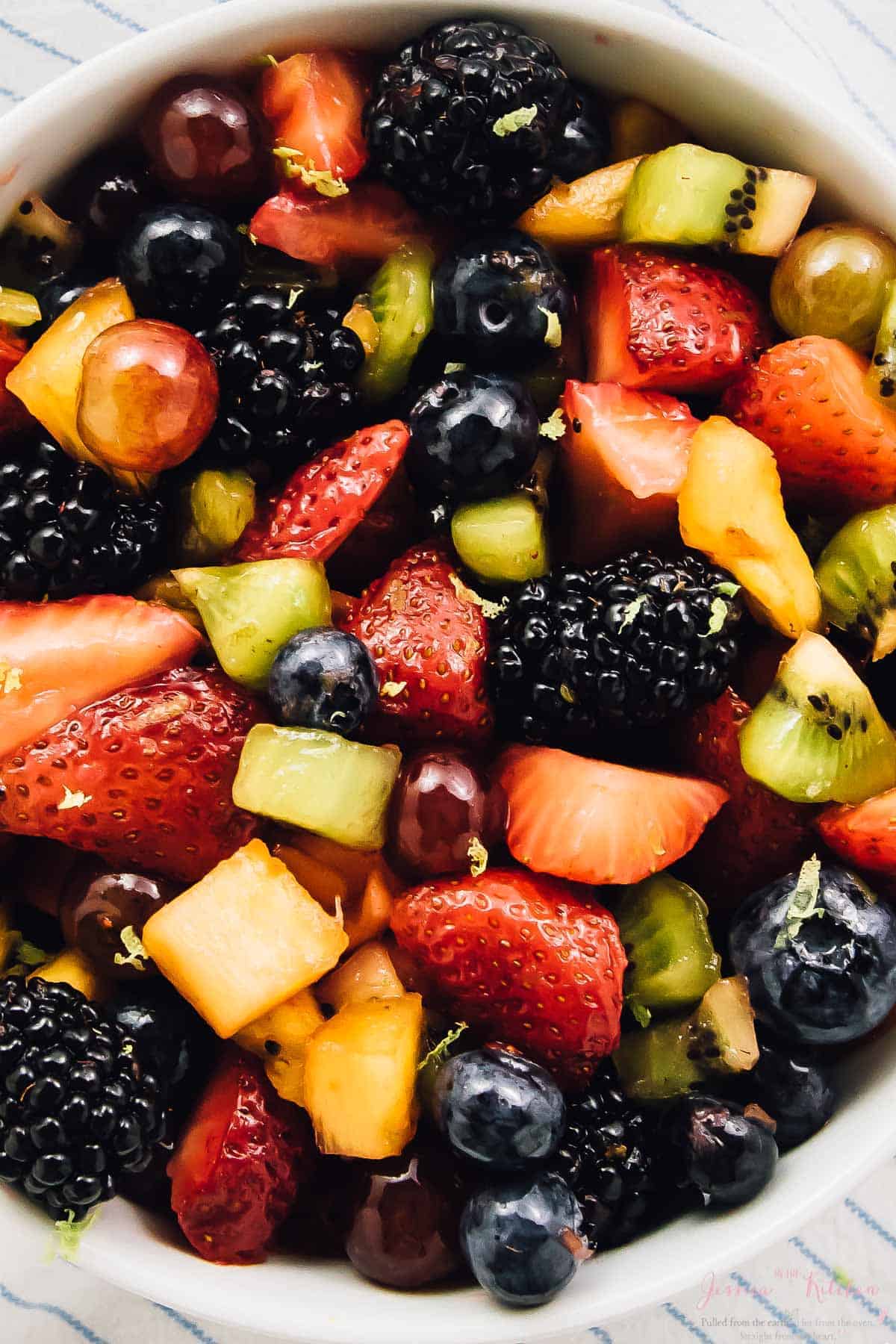 Overhead view of fruit salad in a white bowl on a striped cloth. 