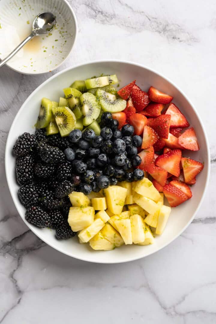 This bright rainbow fruit salad is tossed in a fresh maple lime dressing for a side that's juicy, refreshing, and perfect for any occasion! Blueberries, blackberries, strawberries, kiwi, and pineapple in bowl before being stirred.
