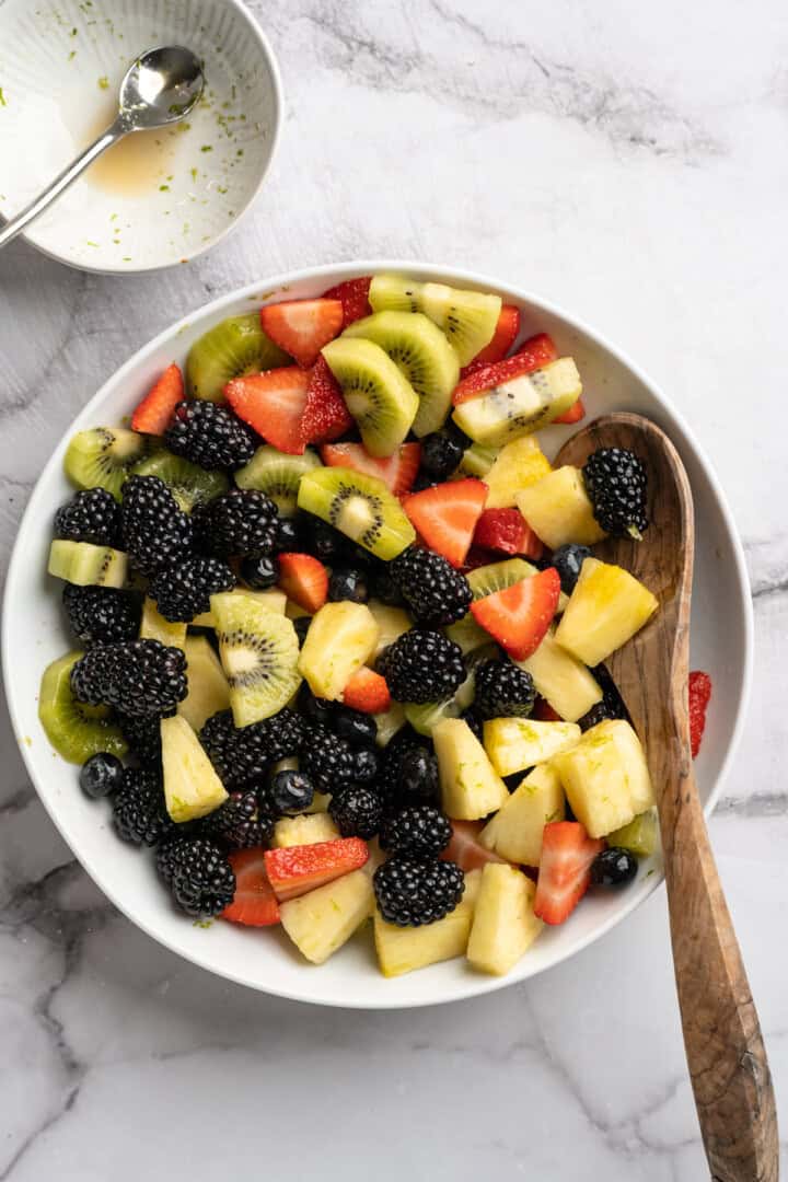 This bright rainbow fruit salad is tossed in a fresh maple lime dressing for a side that's juicy, refreshing, and perfect for any occasion! Berries, kiwi, and pineapple in bowl with wooden spoon.