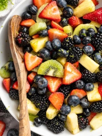 Wooden spoon resting in serving bowl of rainbow fruit salad.